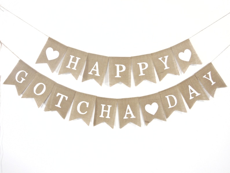 May include: A burlap banner with the text "Happy Gotcha Day" in white letters. The banner has white hearts cut out of it.
