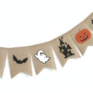 Puede incluir: Una guirnalda de arpillera con dise&ntilde;os de Halloween. Cada bander&iacute;n presenta una imagen diferente: caramelos de ma&iacute;z, un murci&eacute;lago, un fantasma, una casa embrujada, una calabaza y un gato negro. La guirnalda est&aacute; ensartada en una cuerda blanca.