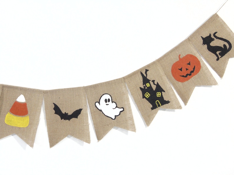 Puede incluir: Una guirnalda de arpillera con dise&ntilde;os de Halloween. Cada bander&iacute;n presenta una imagen diferente: caramelos de ma&iacute;z, un murci&eacute;lago, un fantasma, una casa embrujada, una calabaza y un gato negro. La guirnalda est&aacute; ensartada en una cuerda blanca.