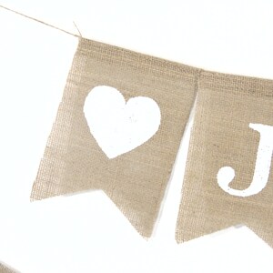 Puede incluir: Una pancarta de arpillera con corazones pintados de blanco y la letra J. La pancarta est&aacute; atada con una cuerda.