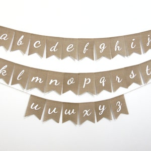 May include: A burlap banner with the alphabet in white cursive letters. The banner is strung on a white background.