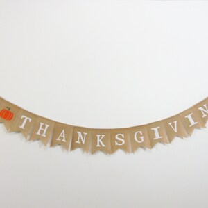 Op de afbeelding: Een banner van jute met de woorden "Happy Thanksgiving" in witte letters. De banner heeft een kleine oranje pompoen met een groene steel in het gedeelte "Happy".