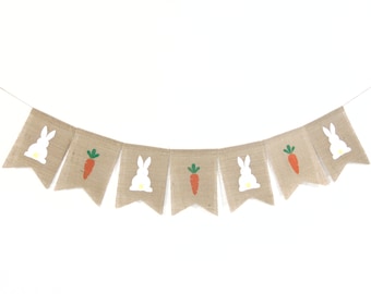 May include: A burlap banner with alternating images of white bunnies and orange carrots with green tops. The banner is strung on a white background.