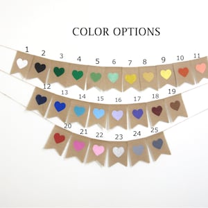 May include: A burlap banner with 25 different coloured heart shapes. The banner is strung with white twine. The colours include white, black, green, teal, yellow, orange, red, pink, blue, purple, brown, and grey.
