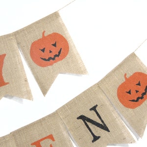 Puede incluir: Una guirnalda de Halloween de arpillera con letras y calabazas naranjas. La guirnalda deletrea "HALLOWEEN" con letras naranjas y caras de calabaza con recortes negros. Est&aacute; ensartada en una fina cuerda de color natural.