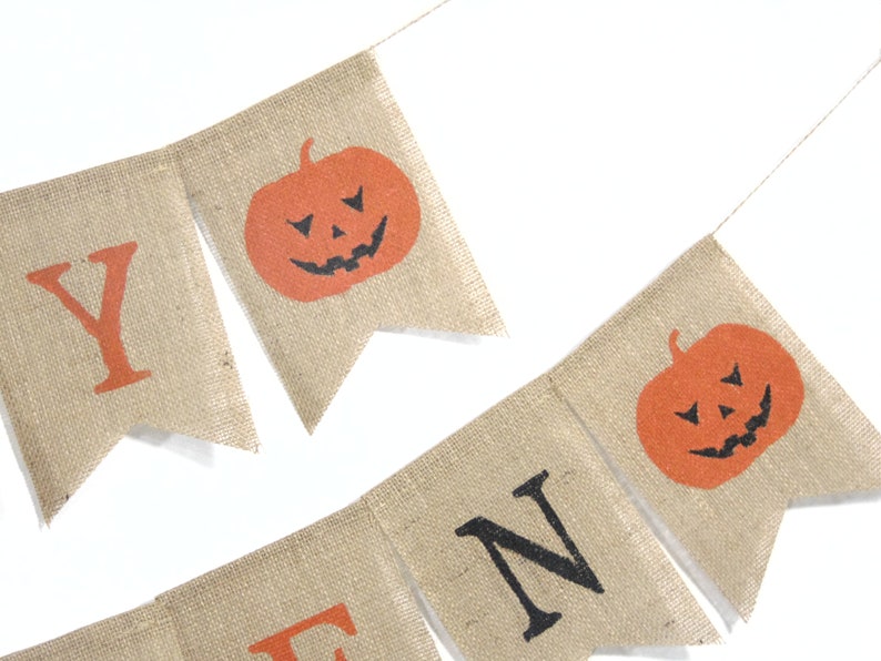 Puede incluir: Una guirnalda de Halloween de arpillera con letras y calabazas naranjas. La guirnalda deletrea "HALLOWEEN" con letras naranjas y caras de calabaza con recortes negros. Est&aacute; ensartada en una fina cuerda de color natural.