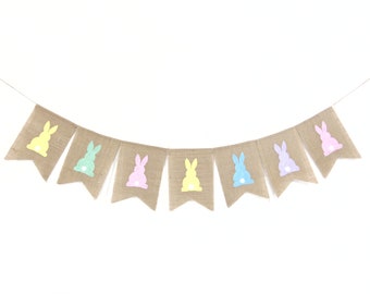 May include: A burlap banner with pastel colored bunny silhouettes. The banner is strung together with twine and has seven flags.