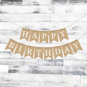 May include: A burlap banner with the words "Happy Birthday" in white letters. The banner is strung on a white string and is hanging against a white wood background.