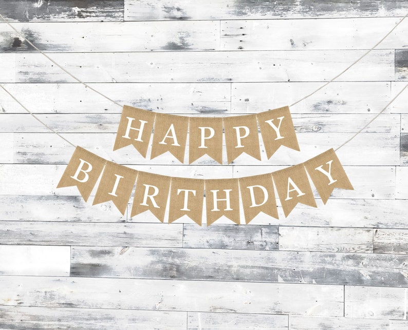 May include: A burlap banner with the words "Happy Birthday" in white letters. The banner is strung on a white string and is hanging against a white wood background.