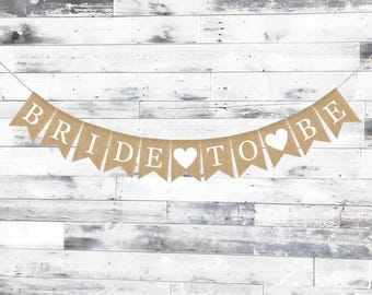 May include: A burlap banner with the words "BRIDE TO BE" in white capital letters. White heart shapes separate the words. The banner is suspended against a white and gray wooden plank background.