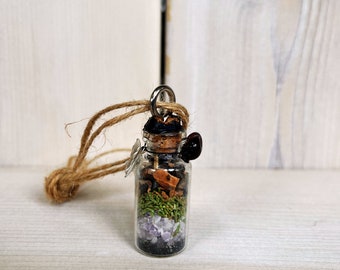 May include: A small glass bottle filled with various herbs, crystals, and other natural elements, suspended from a brown twine cord. The bottle has a cork stopper and is sealed with black wax.
