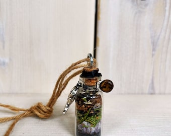 May include: A small glass vial pendant suspended on a brown jute cord. The vial is filled with layers of black sand, purple amethyst crystals, green pine needles, brown soil, and small white stones, sealed with a cork and a metal loop.