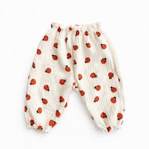 May include: Off-white trousers featuring an all-over print of small red ladybirds with black spots. The trousers have a gathered elastic waistband and elasticated cuffs at the ankles, crafted from a textured, lightweight fabric with a crinkled appearance.