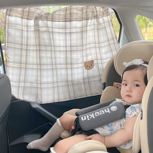 May include: A baby in a car seat with a gray and white plaid sunshade with a bear embroidered on it. The sunshade is attached to the window of the car. The baby is wearing a white and blue floral dress and a white headband. The car seat has a gray strap with the word "meekin" printed on it.