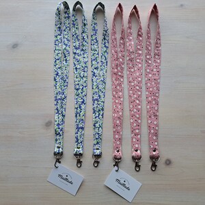 May include: Five fabric lanyards with floral patterns. Two lanyards are blue with white flowers, and three lanyards are pink with white flowers. The lanyards have silver metal clasps.