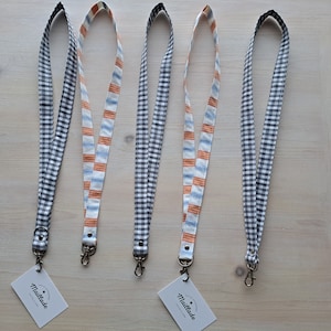 May include: Five fabric lanyards with metal clasps and small white tags. Two lanyards have a black and white gingham pattern, one has orange, white, and blue stripes, and two have the same gingham pattern.