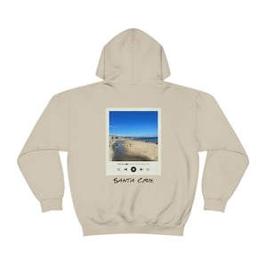 Könnte beinhalten: Ein beiger Hoodie mit einem Foto einer Strandszene auf der Rückseite. Das Foto ist in einem weißen Quadrat mit einem schwarzen Wiedergabetaste und dem Text "SANTA CRUZ" darunter gerahmt.