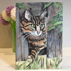 May include: Watercolor painting of a tabby cat with green eyes peeking out from behind a wooden fence. The cat is looking to the right of the image. The background is a watercolor wash of green and brown.