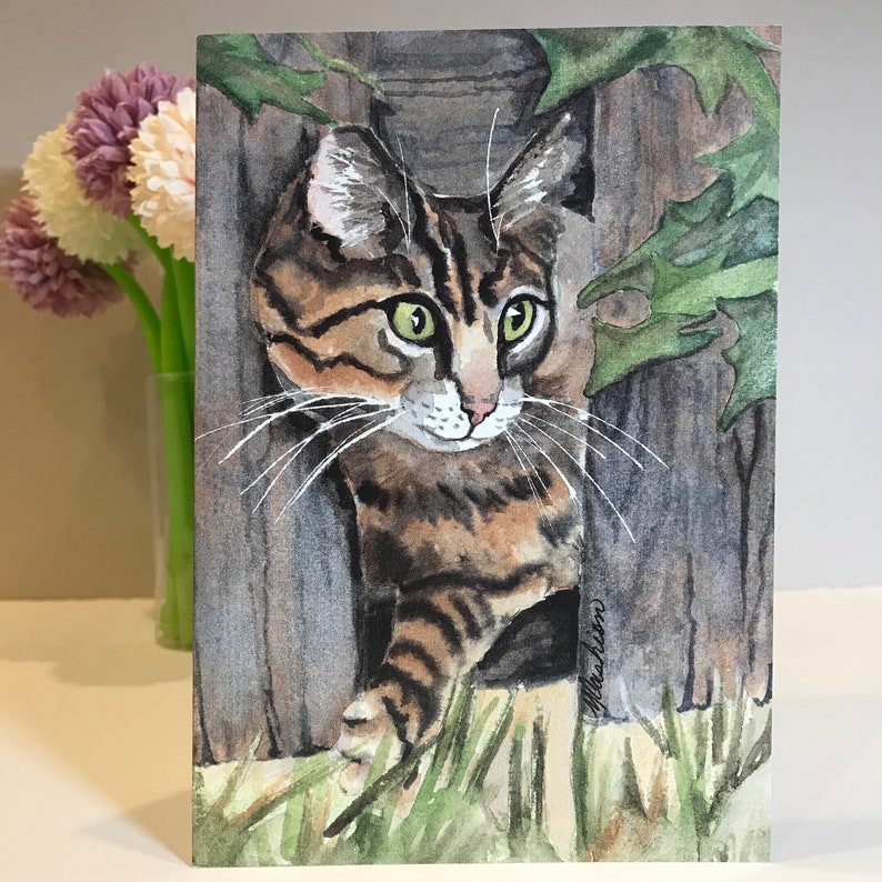 May include: Watercolor painting of a tabby cat with green eyes peeking out from behind a wooden fence. The cat is looking to the right of the image. The background is a watercolor wash of green and brown.