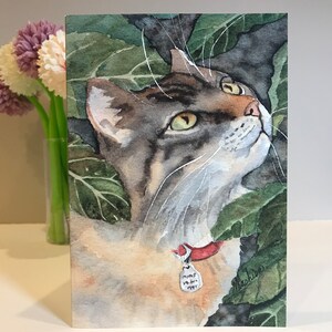 May include: Watercolor painting of a tabby cat with green eyes, wearing a red collar with a tag that says "mas-ter-my", looking up at something off-camera. The cat is surrounded by green leaves.