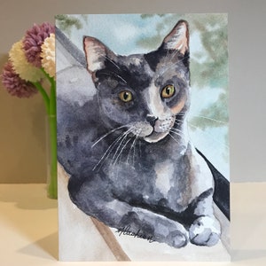 May include: Watercolor painting of a gray and white cat with yellow eyes. The cat is lying down and looking at the viewer. The background is a light blue and green watercolor wash.