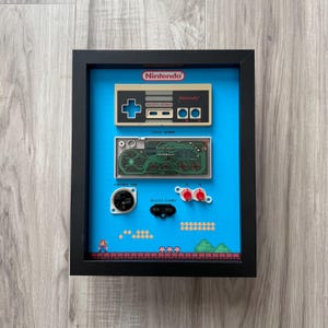 May include: Framed artwork featuring a Nintendo controller and circuit board components against a blue background. The design includes a pixelated Mario scene at the bottom. The frame is black, and the Nintendo logo is at the top.