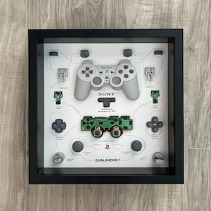 May include: Framed PlayStation DualShock 1 controller parts display. The gray controller and components are arranged on a white background with a black frame. Text includes "SONY" and "Launched on November 20, 1997."