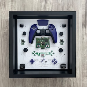 May include: A disassembled PlayStation DualSense wireless controller in a black frame. The controller is shown in its component parts, including the circuit boards, buttons, and vibration motors. The text "DUALSENSE WIRELESS CONTROLLER" and "HEIGHTEN YOUR SENSES" is printed on the white background.