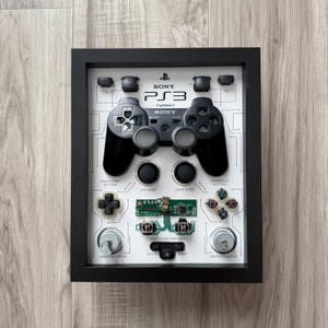 May include: Framed artwork featuring a deconstructed PlayStation 3 controller. The black frame surrounds a white background with the controller's components, including buttons, sticks, and circuit board. The Sony and PS3 logos are visible.