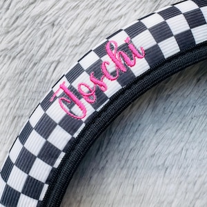 Puede incluir: Funda de volante a cuadros blancos y negros con el nombre "Jeschi" bordado en rosa en cursiva. La funda tiene un ribete negro y está sobre un fondo gris suave.