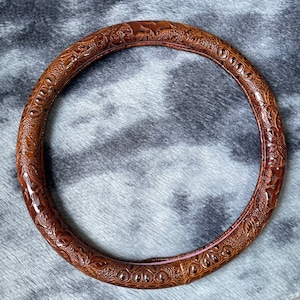 May include: A brown, circular steering wheel cover with an embossed floral and geometric pattern. The cover is made of a textured material and has a glossy finish. The interior edge has a pink accent.