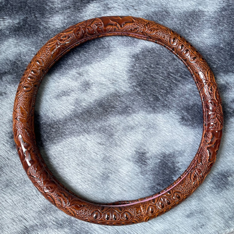 May include: A brown, circular steering wheel cover with an embossed floral and geometric pattern. The cover is made of a textured material and has a glossy finish. The interior edge has a pink accent.