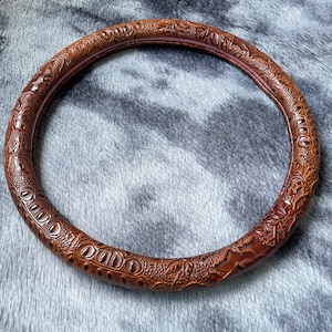 May include: A brown, circular steering wheel cover with a textured, crocodile-like pattern. The cover has a dark brown exterior and a lighter brown interior, with a hint of pink. The design features embossed details.