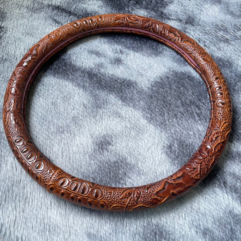 May include: A brown, circular steering wheel cover with a textured, crocodile-like pattern. The cover has a dark brown exterior and a lighter brown interior, with a hint of pink. The design features embossed details.