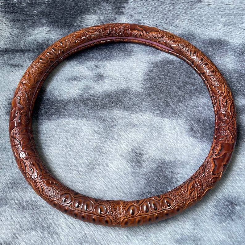May include: A brown, intricately carved steering wheel cover. The cover features a detailed embossed pattern with a crocodile or alligator skin design. The interior rim has a pinkish-brown hue.