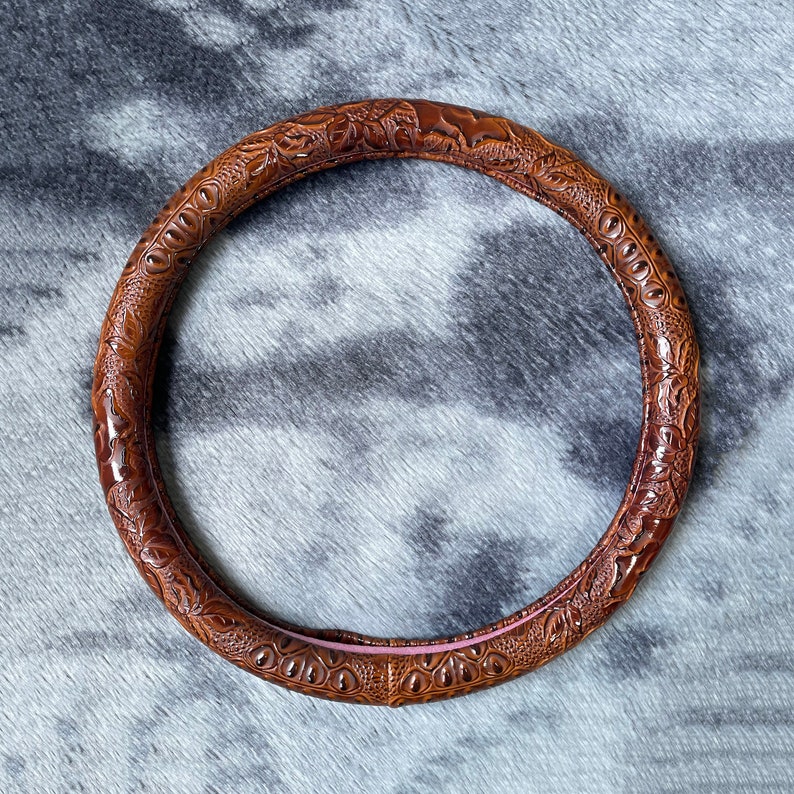 May include: Brown steering wheel cover with an embossed crocodile skin pattern. The cover is circular and has a glossy finish. The interior is lined with a pink material. The cover is designed to fit around a steering wheel.