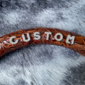 May include: A brown leather steering wheel cover with the word "CUSTOM" spelled out in silver rhinestone letters. The cover has a detailed floral pattern and is set against a gray, furry background.