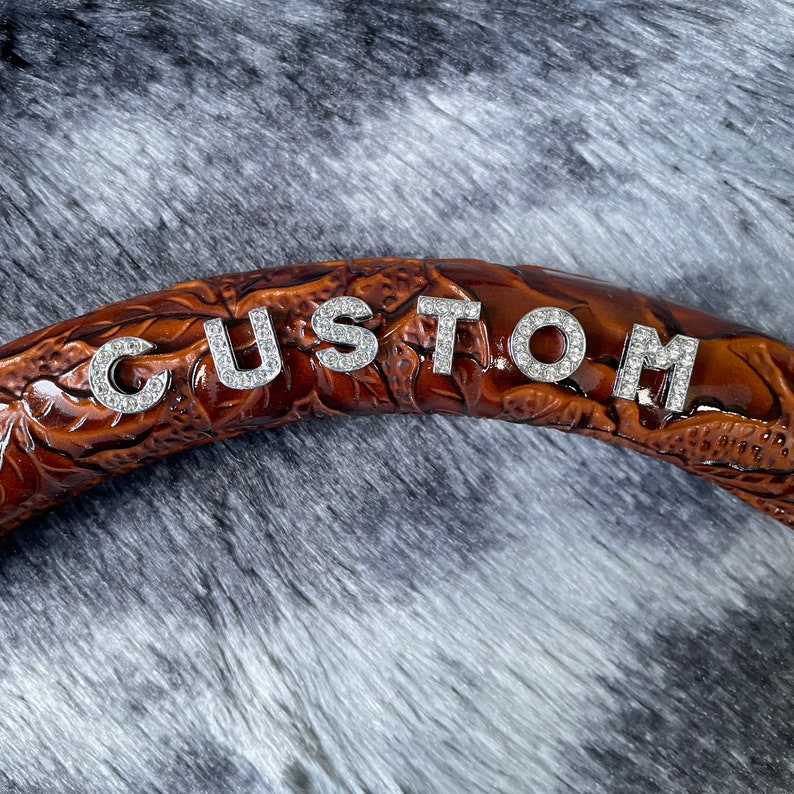May include: A brown leather steering wheel cover with the word "CUSTOM" spelled out in silver rhinestone letters. The cover has a detailed floral pattern and is set against a gray, furry background.