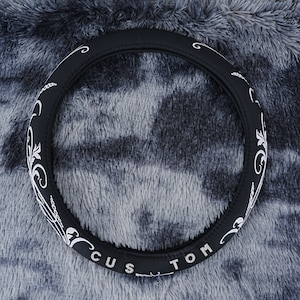 May include: Black steering wheel cover with white embroidery. The embroidery features a floral design and the word "CUSTOM".