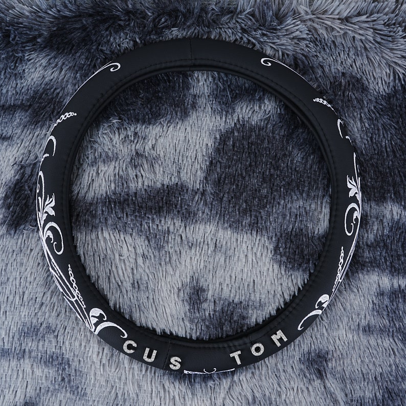 May include: Black steering wheel cover with white embroidery. The embroidery features a floral design and the word "CUSTOM".