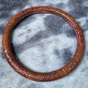 May include: A brown, circular steering wheel cover with an embossed, textured pattern. The cover features a detailed design with floral and reptile-like motifs. The interior edge has a thin, reddish-brown accent. The cover is designed to fit around a steering wheel.