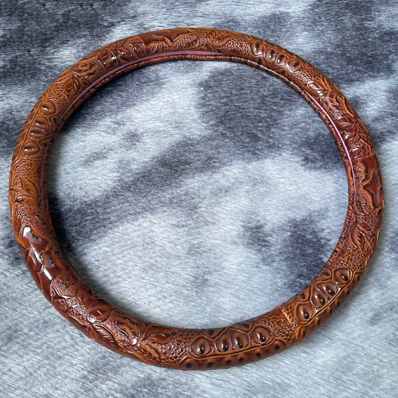 May include: A brown, circular steering wheel cover with an embossed, textured pattern. The cover features a detailed design with floral and reptile-like motifs. The interior edge has a thin, reddish-brown accent. The cover is designed to fit around a steering wheel.
