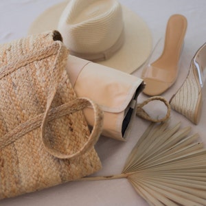 Könnte beinhalten: Eine Strohtasche, ein Strohhut, ein Paar transparente Sandalen mit Absatz und ein Fächer aus Palmblatt. Die Gegenstände sind auf einer weißen Oberfläche angeordnet.