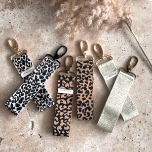 Può includere: Quattro portachiavi con anelli dorati e cinturini in tessuto. Due cinturini sono a stampa leopardata, uno è marrone chiaro con un luccichio e uno è beige chiaro con un luccichio. I cinturini hanno la scritta "ANNIKA" stampata.