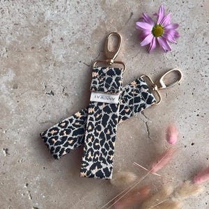 May include: Two gold keychains with leopard print fabric straps. The straps have the word "ANNWAY" printed on them. The keychains are on a grey textured surface.