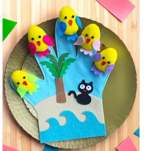 May include: A felt hand puppet depicting a beach scene with a blue ocean, palm tree, and a black cat. Five yellow bird finger puppets with colourful wings are on the fingers. The puppet is on a gold plate.