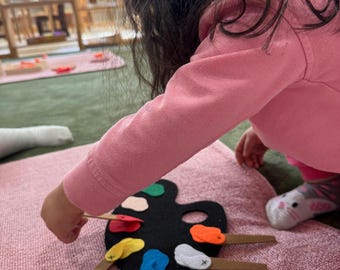 May include: A child's felt art palette with colorful paint blobs and attached felt paintbrushes. The palette is black, and the paint colors include red, yellow, blue, orange, and white. The child is wearing a pink sweatshirt.