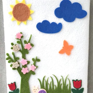 May include: A white felt board decorated with colorful felt cutouts. The scene includes a yellow and brown sun, blue clouds, a tree with pink and glittery flowers, green grass, a snail, an orange butterfly, and red tulips.