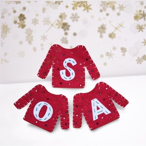 Könnte beinhalten: Rote Filz-Pullover-Ornamente, die das Wort "SOA" mit weißen Buchstaben buchstabieren. Jedes Ornament ist mit kleinen, glänzenden Akzenten verziert. Der Hintergrund zeigt ein festliches Design mit goldenen und weißen Schneeflocken.