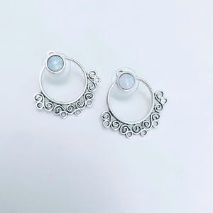 Puede incluir: Un par de pendientes de botón plateados. Cada pendiente presenta un diseño circular con una pequeña piedra de color claro en el centro. El borde exterior del círculo está adornado con un patrón decorativo en espiral.